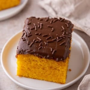 A single square of carrot cake with chocolate frosting and sprinkles on a small white plate