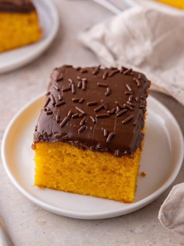 A single square of carrot cake with chocolate frosting and sprinkles on a small white plate