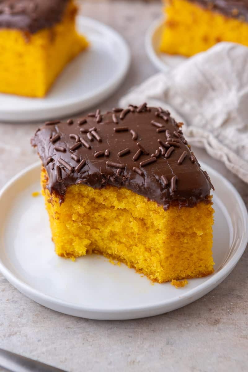 A square slice of Brazilian carrot cake with chocolate glaze on a white plate with more slices in the background