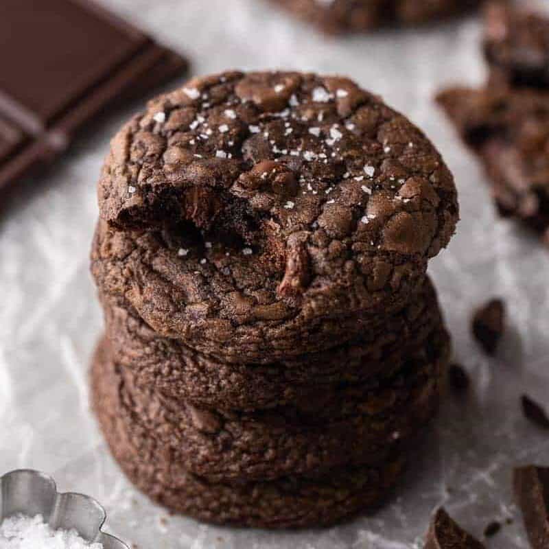 Stack of four Crumbl brownie batter cookies with crinkly tops and flaky sea salt next to dark chocolate squares and chocolate shavings