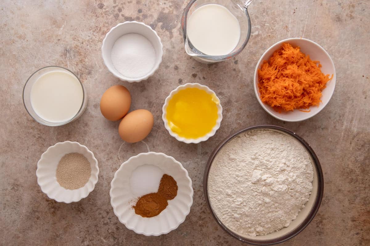 Ingredients for cinnamon roll dough including flour, shredded carrots, eggs, milk, melted butter, sugar, yeast, and spices in small bowls