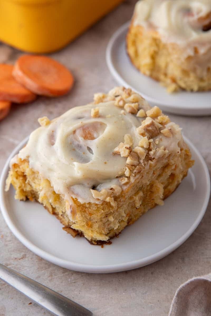 A frosted carrot cake cinnamon roll topped with chopped walnuts on a white plate with sliced carrots and a yellow baking pan in the background
