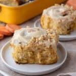 Two carrot cake cinnamon rolls with brown butter cream cheese frosting and chopped walnuts on white plates with a yellow baking pan in the background