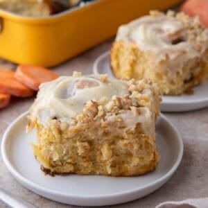 Two carrot cake cinnamon rolls with brown butter cream cheese frosting and chopped walnuts on white plates with a yellow baking pan in the background