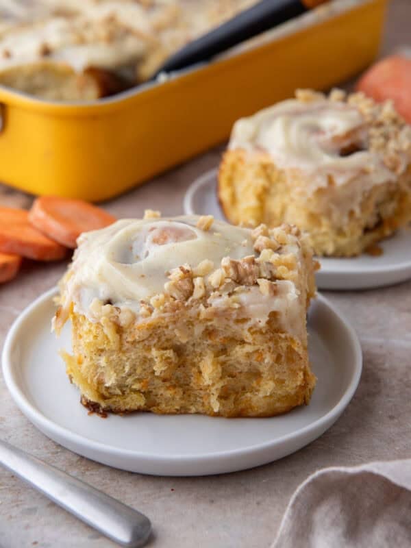 Two carrot cake cinnamon rolls with brown butter cream cheese frosting and chopped walnuts on white plates with a yellow baking pan in the background