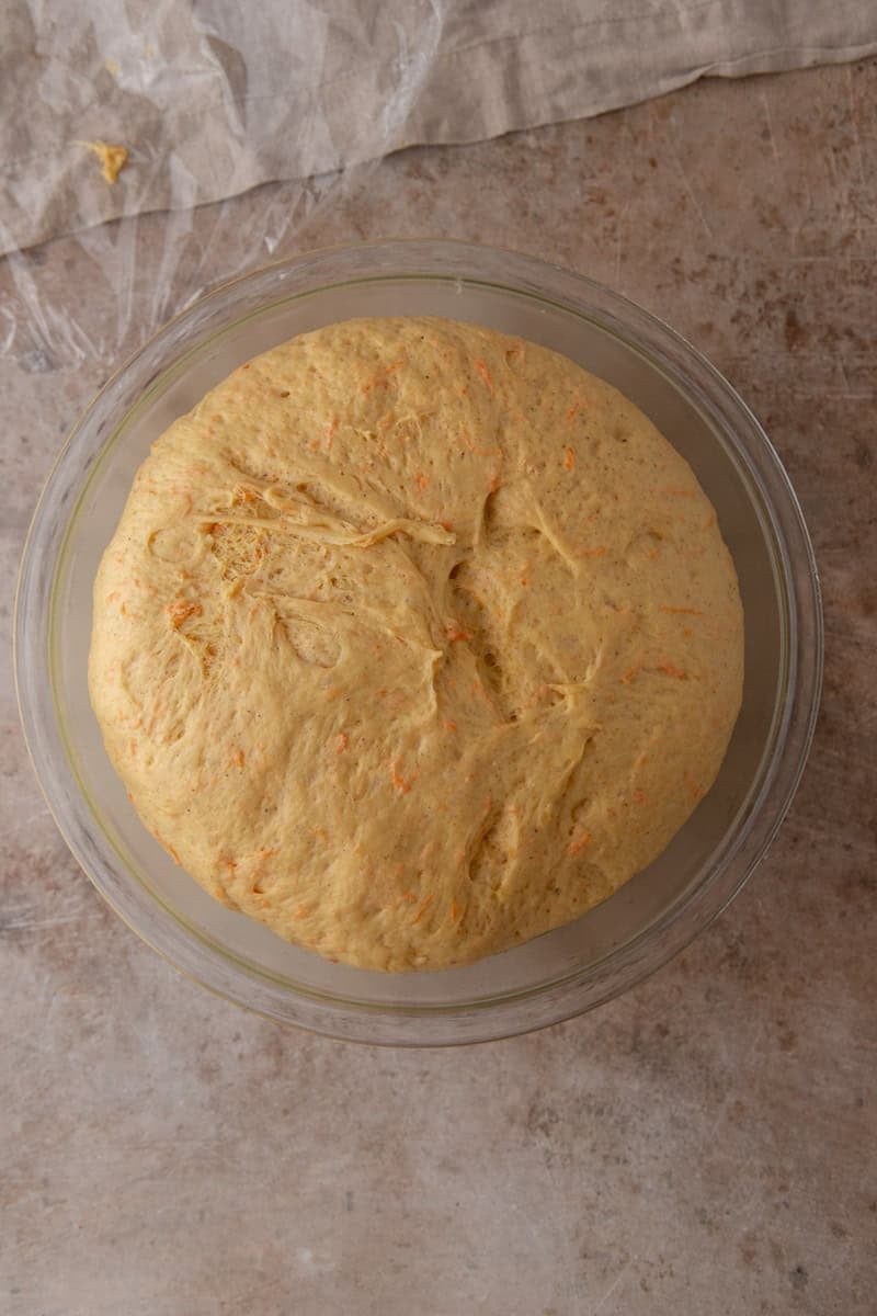 Cinnamon roll dough doubled in size after rising in a glass bowl, with orange carrot flecks visible