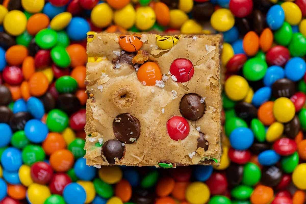 Single M&M chocolate chip blondie bar surrounded by colorful M&M candies showing the chewy texture and baked-in candy pieces