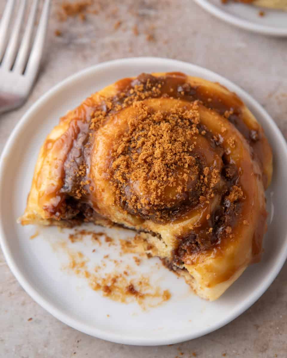 Glazed Biscoff cinnamon roll on a white plate with a bite taken and crushed cookies on top