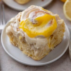 A single lemon roll with lemon curd on a small white plate with lemons in the background