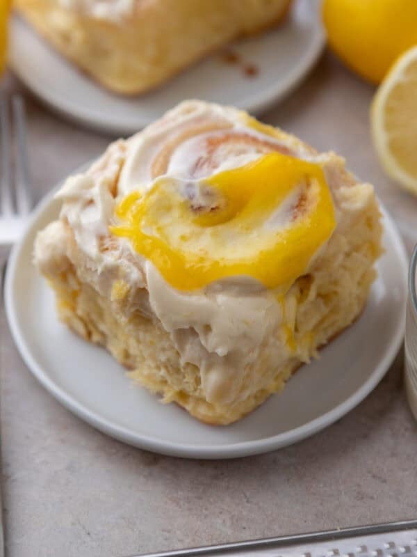 A single lemon roll with lemon curd on a small white plate with lemons in the background