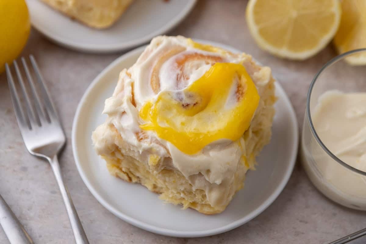 Lemon roll with lemon curd and cream cheese frosting on a white plate with lemons and a glass of frosting nearby