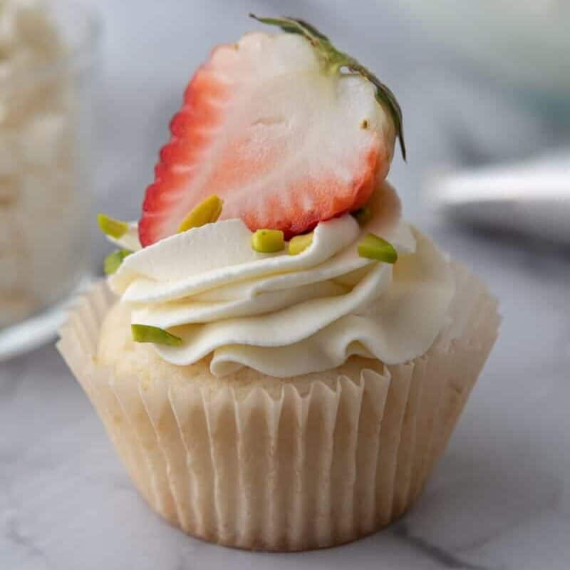 Vanilla cupcake topped with mascarpone frosting, a fresh strawberry slice, and chopped pistachios