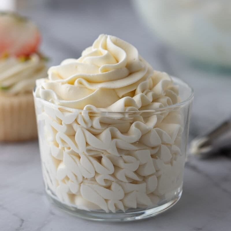Mascarpone frosting recipe piped into a glass bowl with a frosted cupcake in the background