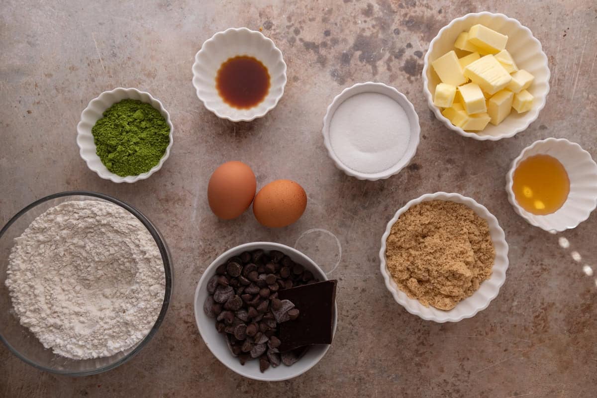 Ingredients for matcha cookies measured out in white bowls including flour, matcha powder, butter, sugars, eggs, chocolate chips, honey, and vanilla