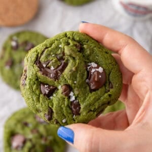 Hand holding a thick green matcha cookie with melty chocolate chunks and flaky sea salt on top