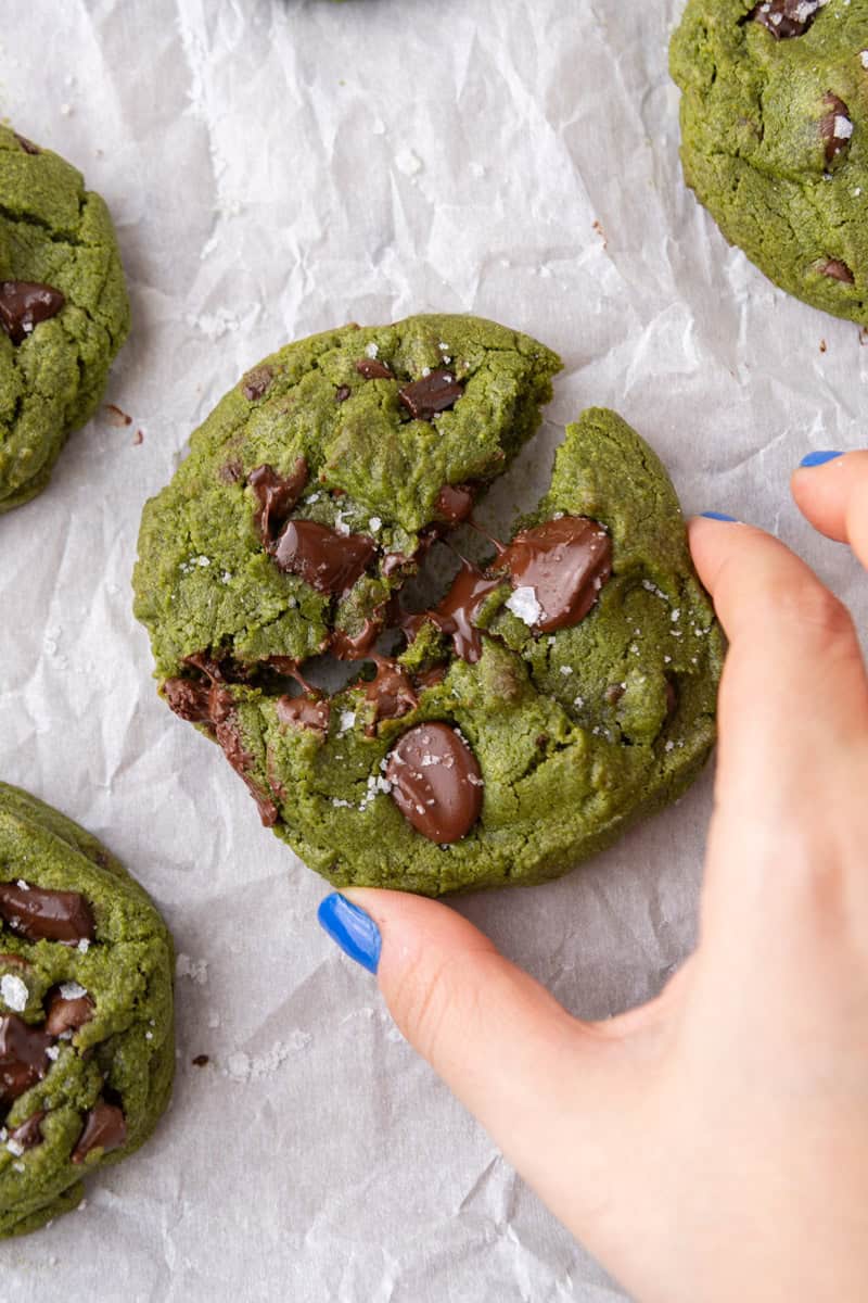 Hand breaking apart a thick chewy matcha cookie with melted chocolate and sea salt flakes on parchment paper