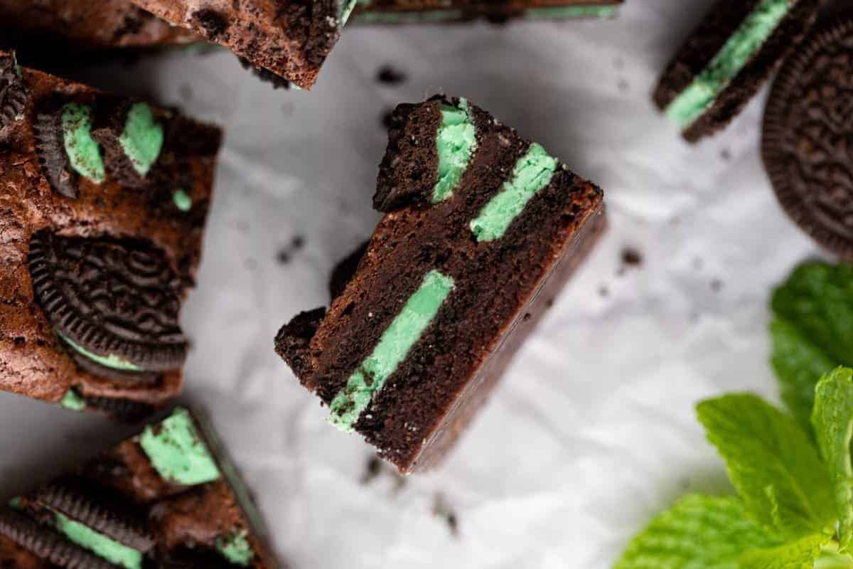 Mint Oreo brownie tilted to show the cross-section with layers of bright green mint Oreo filling baked into the fudgy chocolate brownie surrounded by mint leaves and mint Oreo cookies