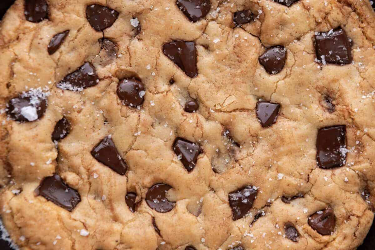 Close-up of the baked surface of a thick skillet cookie showing melted chocolate pools, a golden crackly top, and flaky sea salt crystals