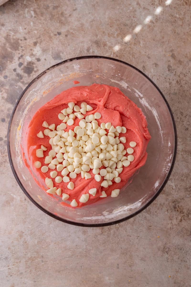 Bright pink strawberry cookie dough in a glass bowl with white chocolate chips on top ready to be folded in