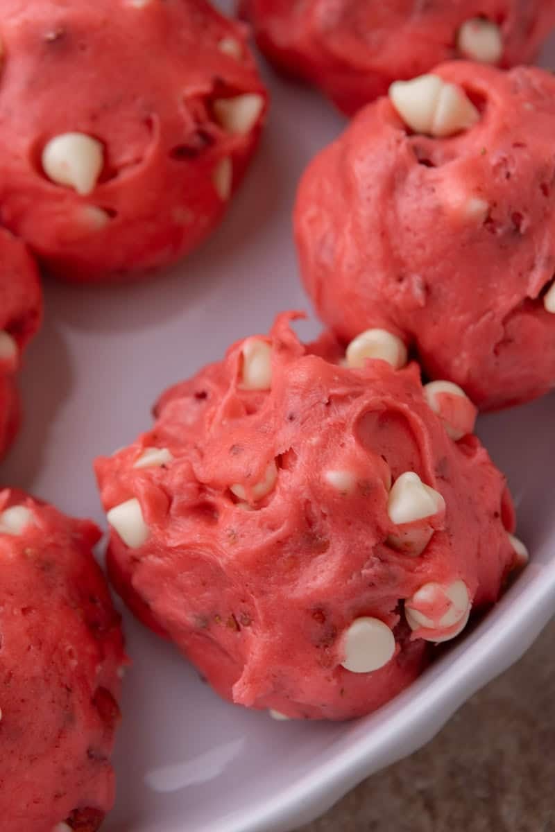 Pink strawberry cookie dough balls with white chocolate chips on a white plate before baking