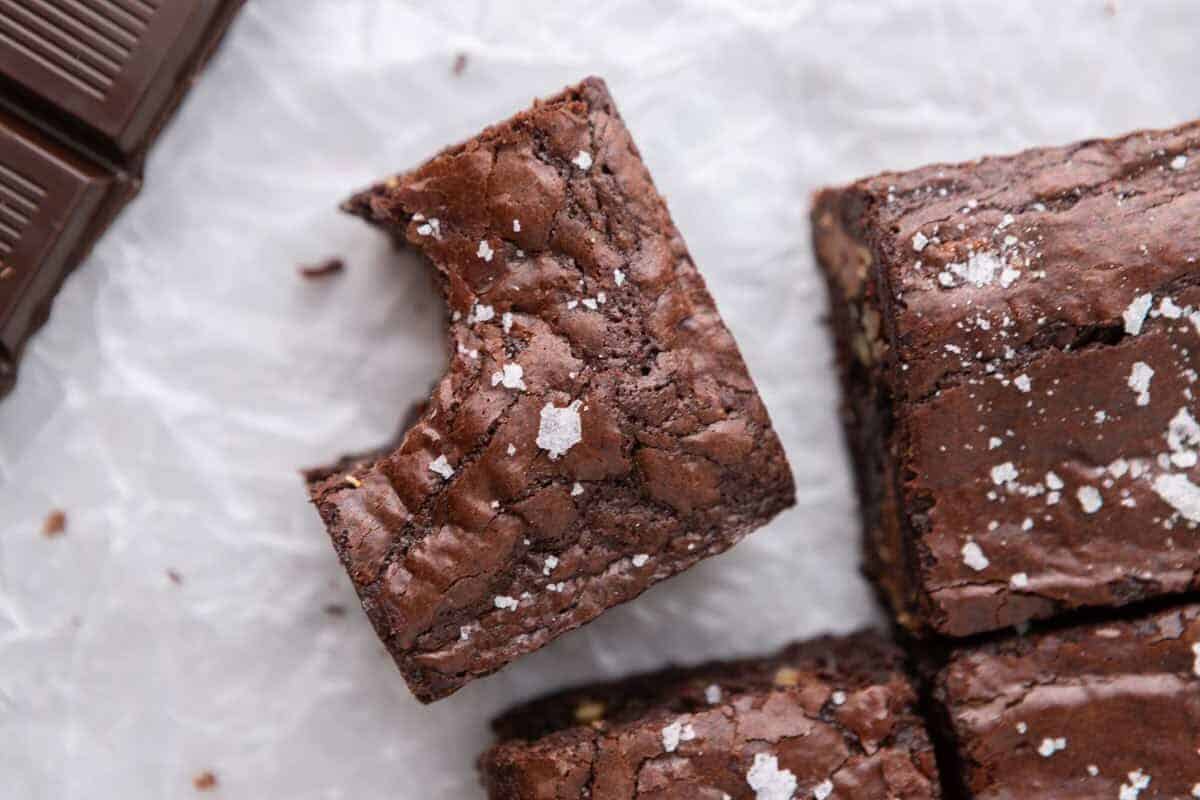 Overhead view of fudgy homemade brownies with crinkly tops and flaky sea salt on parchment paper next to dark chocolate squares