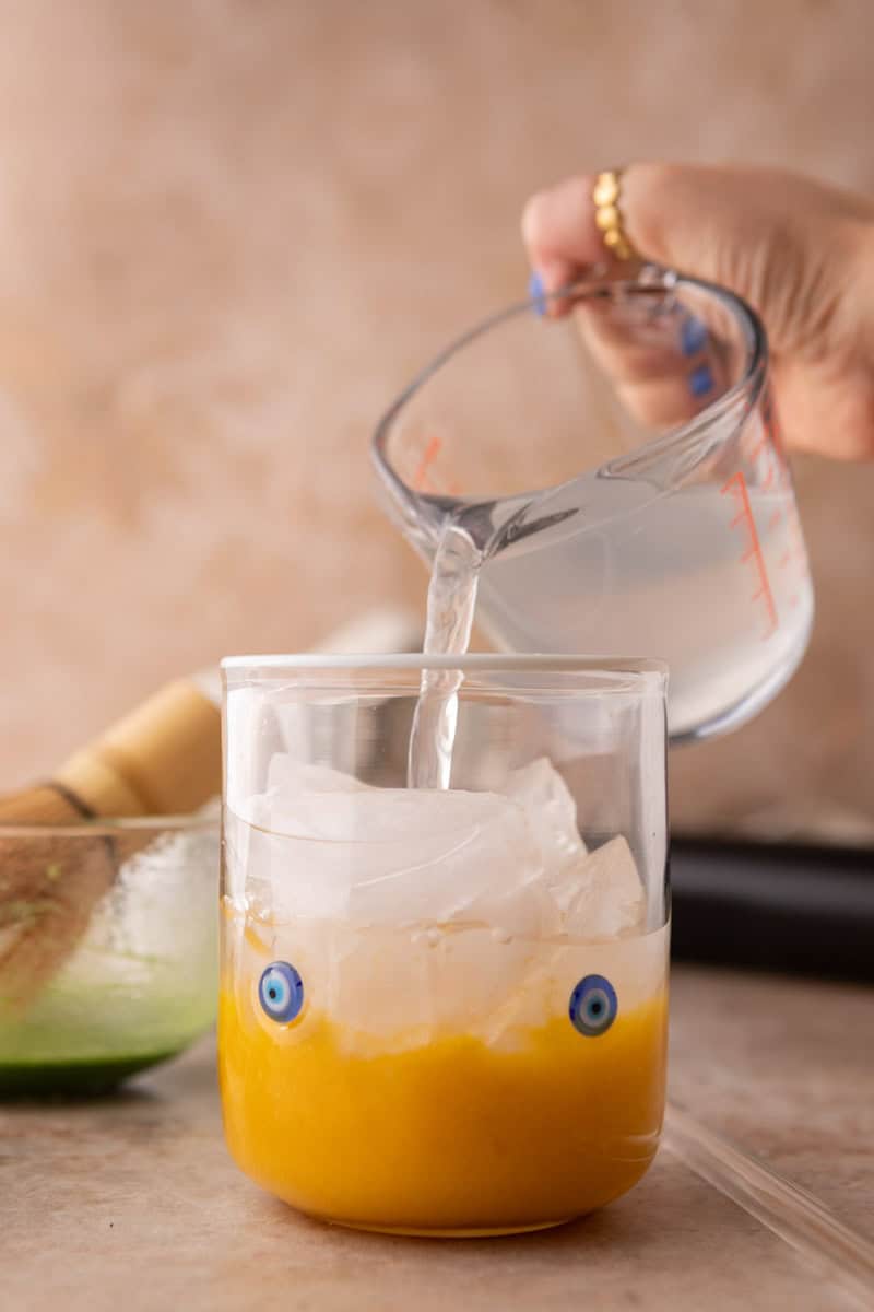 pouring coconut water over ice into a glass with mango puree for mango matcha drink