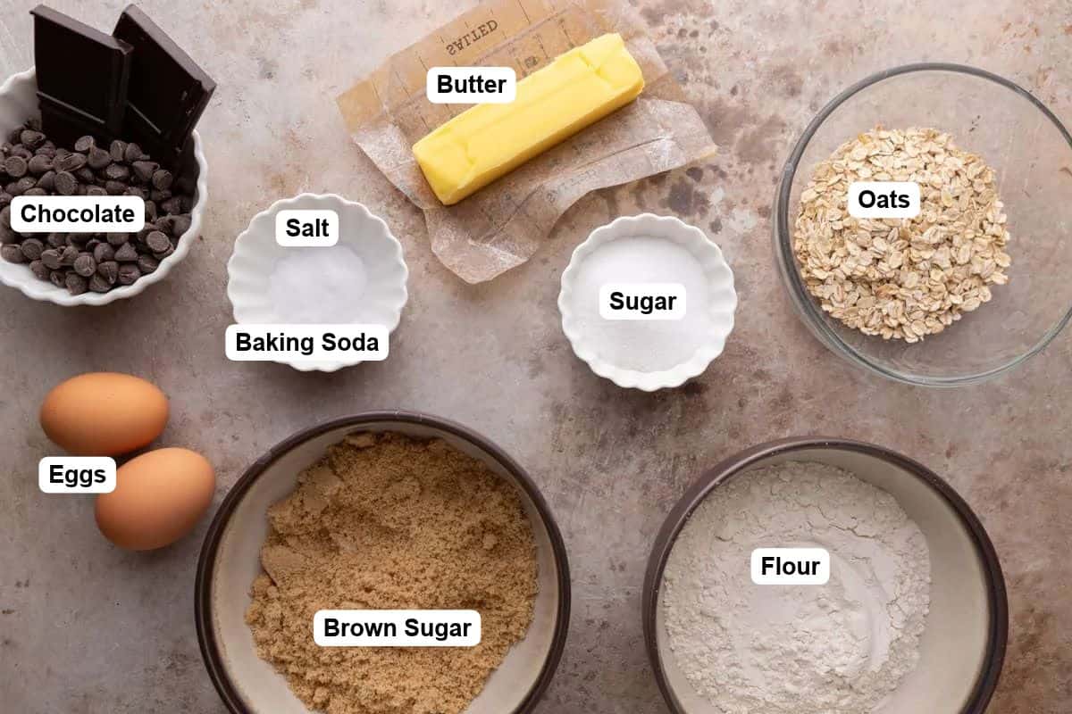 Overhead view of cookie bar ingredients in bowls, including butter, brown sugar, granulated sugar, flour, rolled oats, eggs, chocolate chips, and dark chocolate.