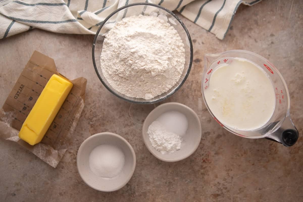 Overhead view of all butter swim biscuit ingredients laid out on a stone surface including a stick of unsalted butter on its wrapper, a glass bowl of flour, two small bowls of sugar and baking powder, and a glass measuring cup of buttermilk.