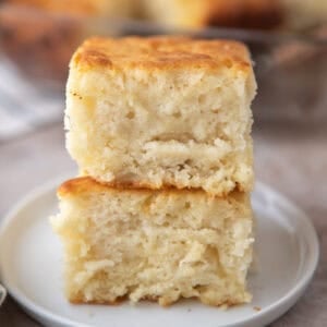 Two butter swim biscuits stacked on a small white plate showing their golden brown tops and soft fluffy interior with the rest of the batch visible in a glass baking dish in the background.