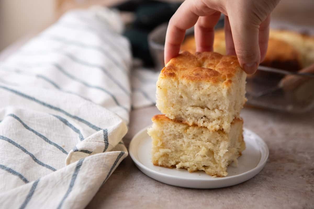 A hand reaching for one of two stacked butter swim biscuits on a small white plate with a blue and white striped kitchen towel and the glass baking dish of biscuits in the background.