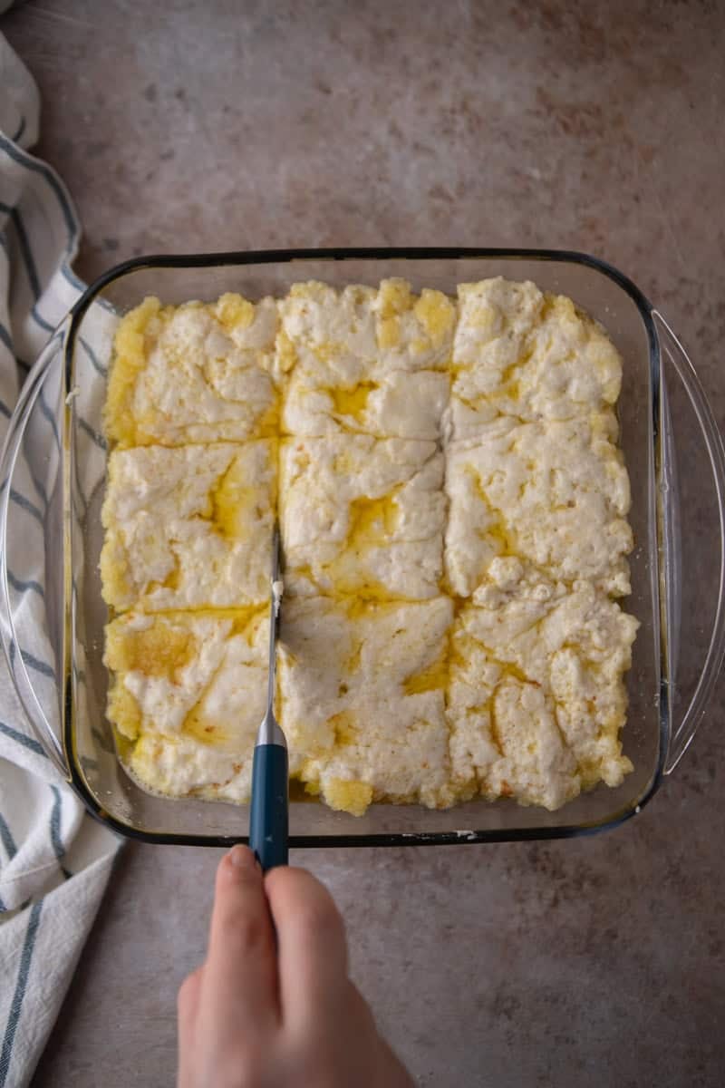 A hand using a knife with a blue handle to score biscuit batter into 9 squares in a 3x3 pattern in a glass 8x8 baking dish with melted butter visible around the cuts.