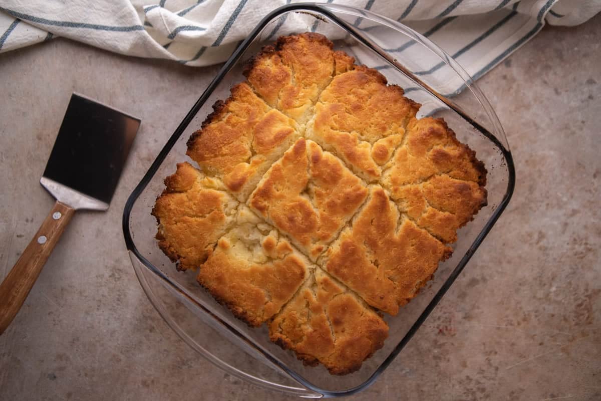 Overhead view of freshly baked butter swim biscuits in a glass 8x8 baking dish with deep golden brown tops, crispy caramelized edges, and visible score lines separating 9 puffy squares.