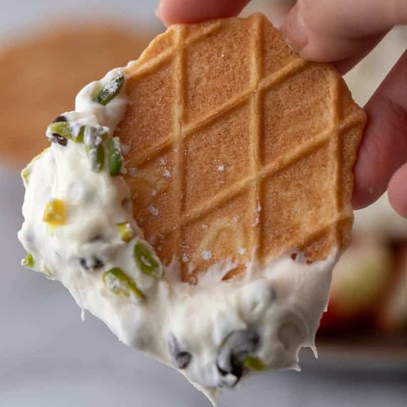 A pizzelle cookie being coated in dip with, showing the irresistible creamy texture and chocolate chip-studded dip clinging to the crisp vanilla wafer.