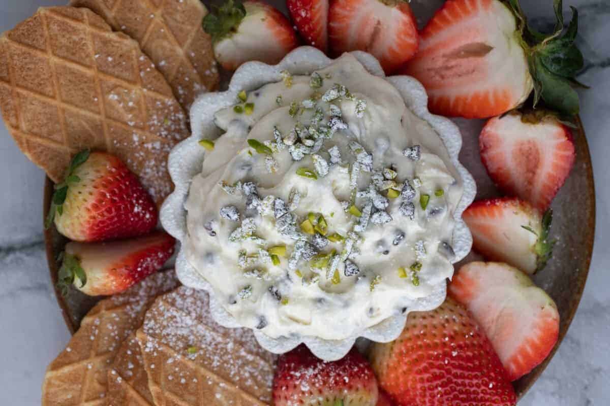Creamy cannoli dip surrounded by fresh strawberries, cookies, and pizzelle. The fluffy ricotta and mascarpone dip is topped with mini chocolate chips and pistachios.