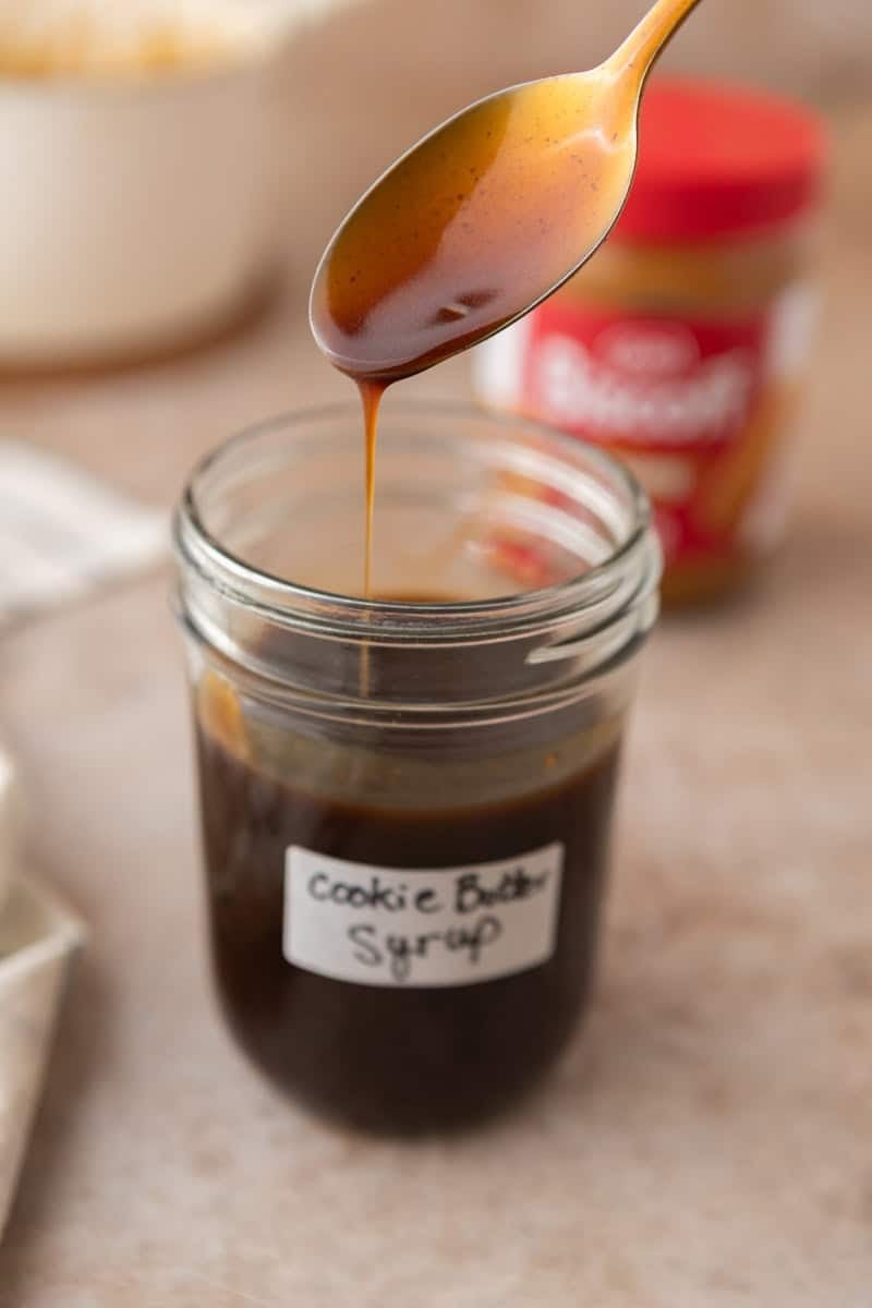 A spoon drizzling homemade cookie butter syrup into a labeled mason jar showing the thick, pourable consistency.