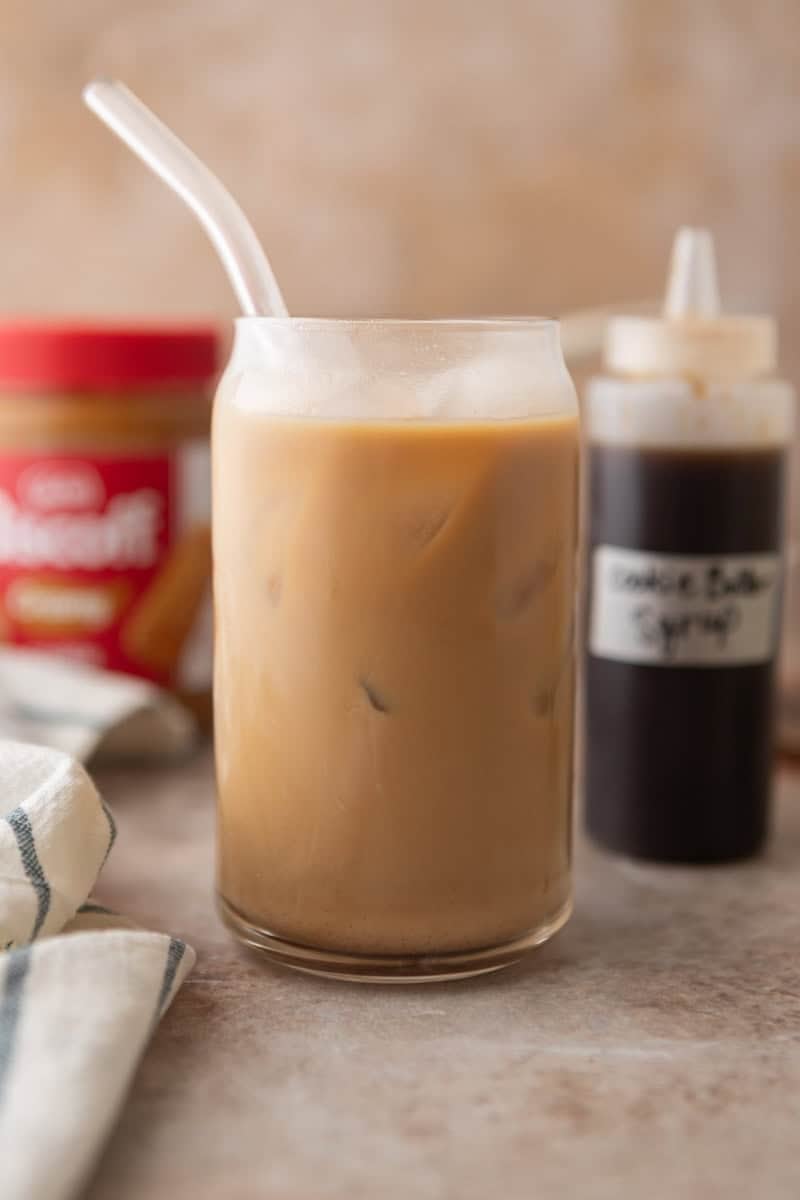 A cookie butter iced latte made with homemade Biscoff syrup in a glass with a straw, with the syrup bottle and Biscoff jar in the background.