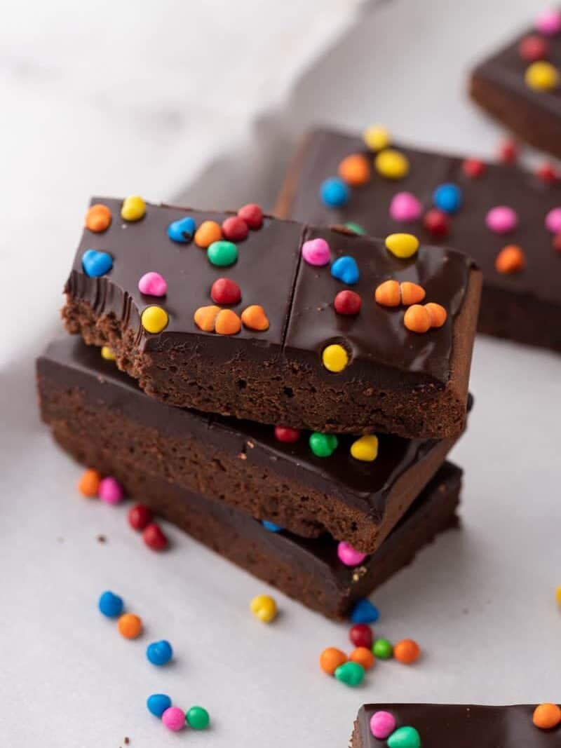 Stack of three homemade cosmic brownies with thick chocolate ganache and colorful rainbow candy chips on top with more candy scattered around