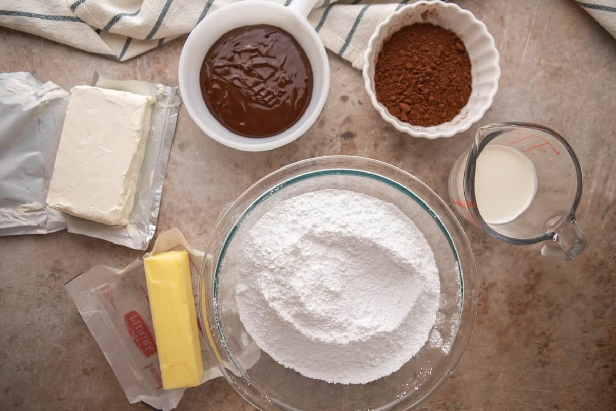 Ingredients for chocolate cream cheese frosting including cream cheese, butter, powdered sugar, cocoa powder, melted chocolate, and heavy cream