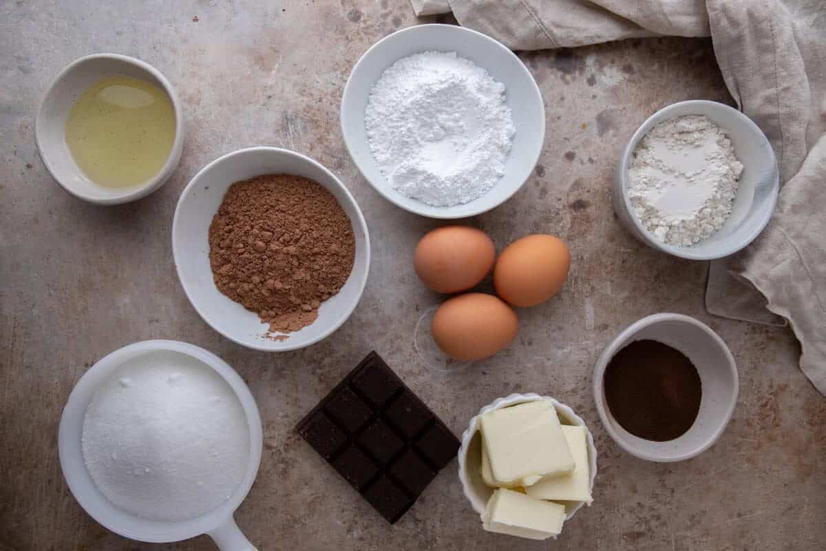 All ingredients for espresso brownies measured in white bowls including cocoa powder, dark chocolate bar, eggs, butter, sugar, and espresso powder