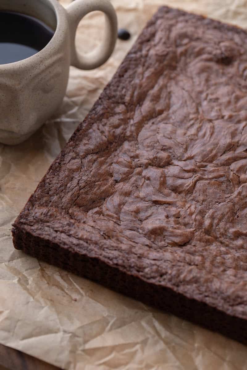 Whole slab of baked brownies with crinkly top on parchment next to a cup of coffee