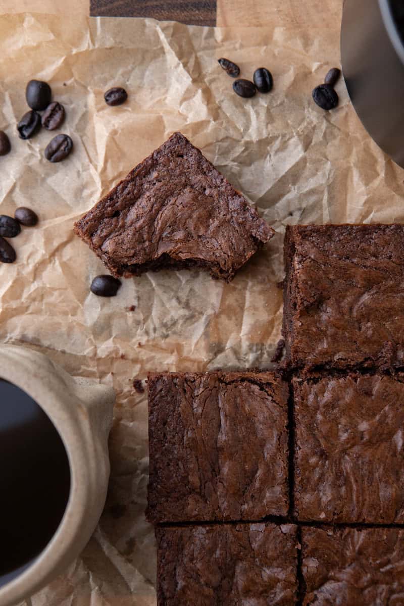 Overhead shot of sliced brownies on parchment with scattered coffee beans and a ceramic mug of espresso