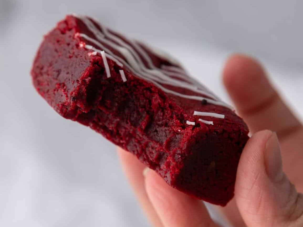 Hand holding a fudgy red velvet brownie with a bite taken out showing the dense vibrant red interior and white chocolate drizzle on top