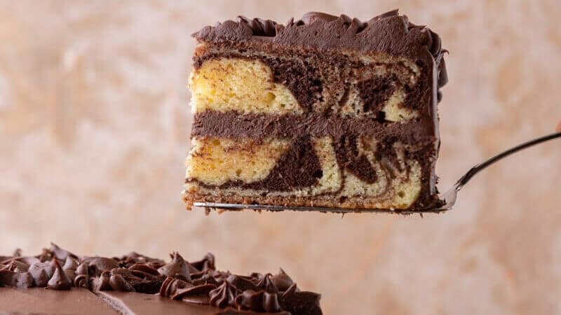 Slice of layered marble cake with chocolate cream cheese frosting lifted on a cake server showing vanilla and chocolate swirls