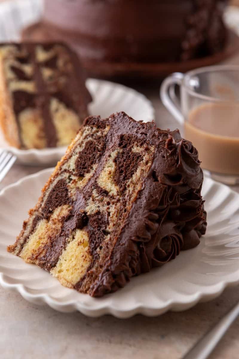 Slice of marble cake with chocolate frosting on a white scalloped plate with coffee in the background