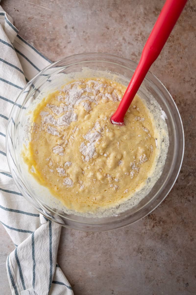 Wet ingredients being folded into dry ingredients with a red spatula in a glass bowl