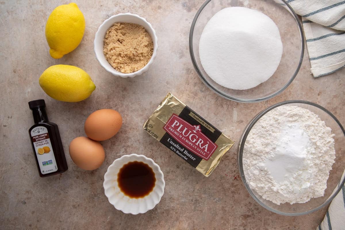 Overhead flatlay of lemon sandwich cookie ingredients including Plugrà butter, lemons, eggs, sugar, flour, and lemon extract.