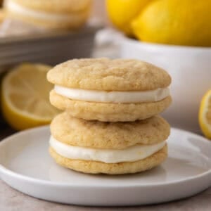 Two lemon sandwich cookies stacked on a small white plate with fresh lemons in the background.