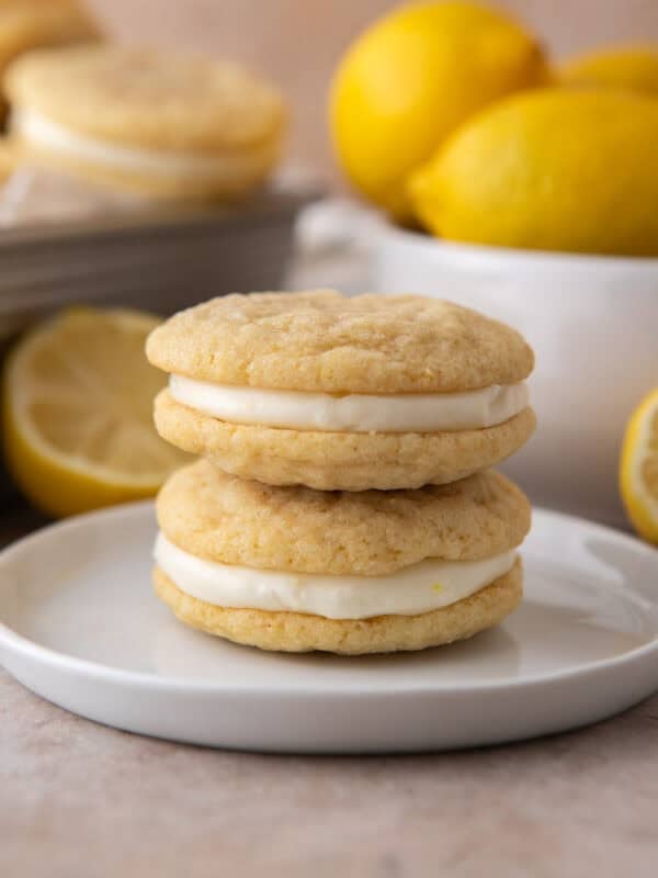 Two lemon sandwich cookies stacked on a small white plate with fresh lemons in the background.