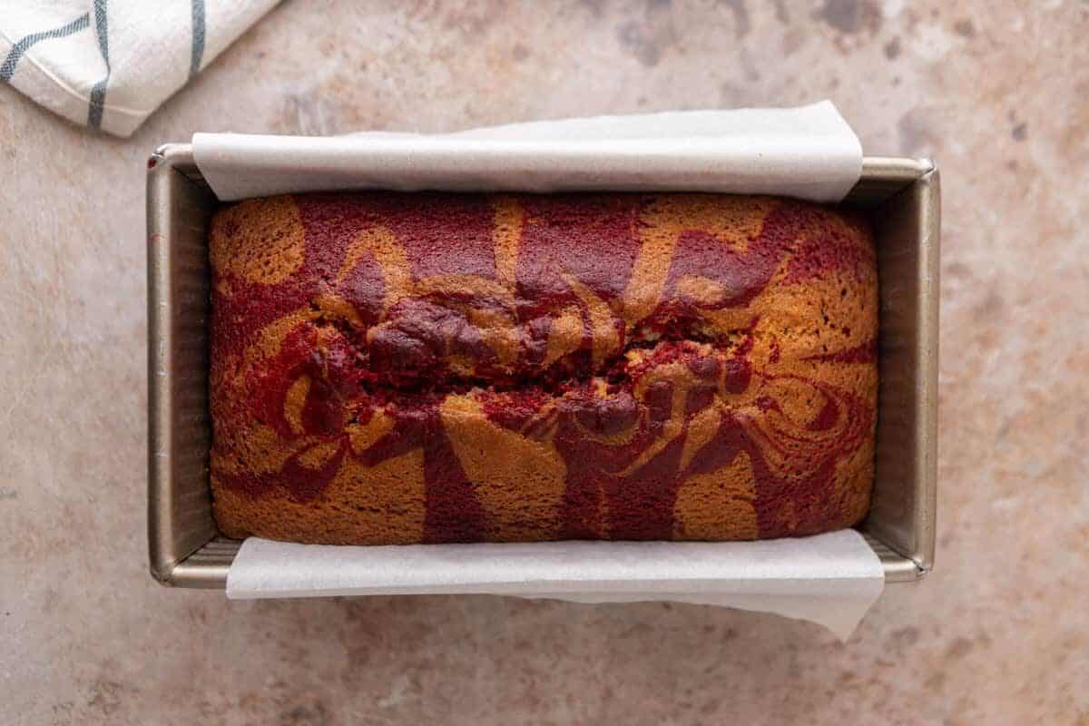 A freshly baked marble loaf cake in a parchment-lined loaf pan with red and golden swirls on top.