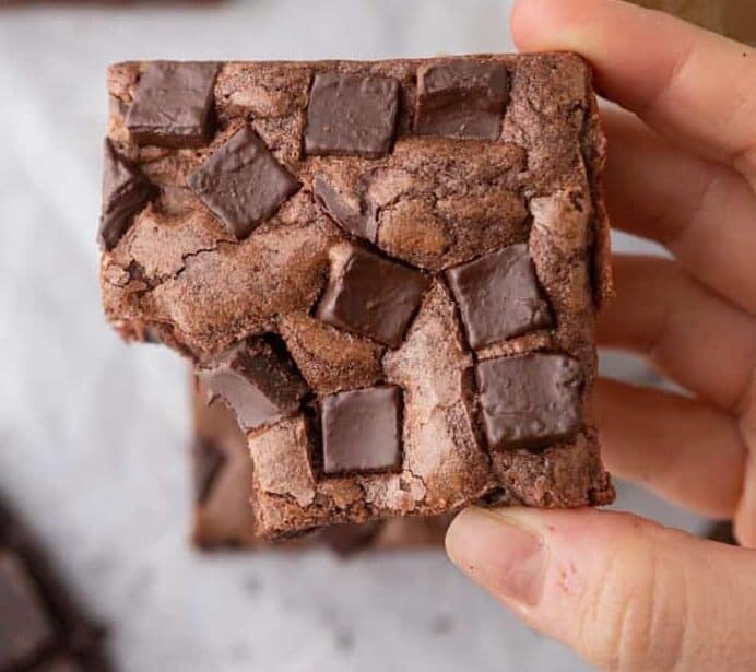 Hand holding a Starbucks copycat double chocolate brownie loaded with chocolate chunks on top with a cup of coffee and more brownies in the background